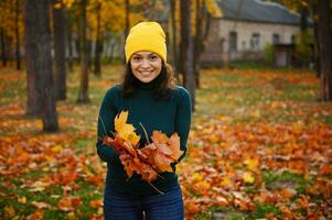 porträtt av en skön leende kvinna i ull- hatt, i de höst parkera, innehav en torr samlade in bukett av färgrik höst gul orange ek och lönn löv och redo för kasta dem upp i de luft foto