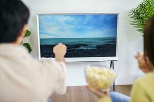 asiatisk familj tittar på smart TV tillsammans och använder sig av avlägsen kontrollant hand innehav popcorn på Hem med de avlägsen kontrollera film eller TV serier utgifterna tid på Hem foto