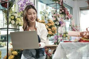 ett ung asiatisk kvinna blomsterhandlare och sme entreprenör i förkläde, Arbetar med bärbar dator, uppkopplad försäljning blommig ordna i färgrik blomma affär Lagra med skön knippa av blommar, och e-handel företag. foto