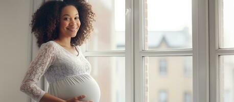 ung gravid svart kvinna leende och ser på tömma Plats njuter henne graviditet nära en fönster på Hem foto