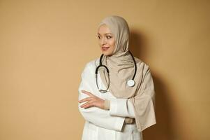 skön lugn muslim läkare kvinna i hijab stående med korsade vapen och ser sida på beige bakgrund med kopia Plats foto