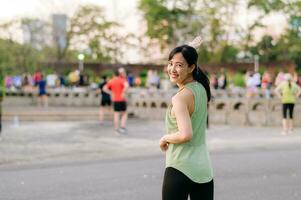 kvinna joggare. passa ung asiatisk kvinna med grön sportkläder aerobics dansa övning i parkera och njuter en friska utomhus. kondition löpare flicka i offentlig parkera. wellness varelse begrepp foto