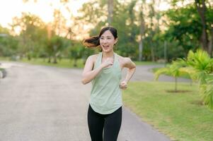 passa asiatisk ung kvinna joggning i parkera leende Lycklig löpning och njuter en friska utomhus- livsstil. kvinna joggare. kondition löpare flicka i offentlig parkera. friska livsstil och wellness varelse begrepp foto