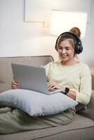 ung skön asiatisk kvinna avkopplande och lyssnande till musik använder sig av hörlurar, hon är liggande i soffa. ung kvinna njuter på Hem använder sig av bärbar dator. foto