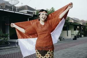 Lycklig leende indonesiska kvinna bär röd kebaya innehav Indonesiens flagga till fira indonesien oberoende dag. utomhus- fotografering begrepp foto