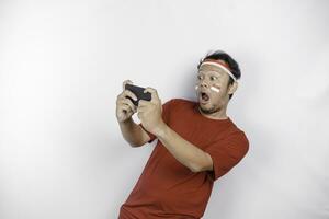 en porträtt av en chockade asiatisk man bär pannband, innehav hans telefon medan hans mun bred öppna, isolerat förbi vit bakgrund. Indonesiens oberoende dag begrepp foto