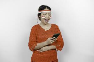 en omtänksam asiatisk kvinna bär röd kebaya och pannband, innehav henne telefon och mage, tänkande handla om Vad till äta eller ordning, isolerat förbi vit bakgrund. Indonesiens oberoende dag foto