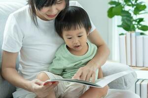 Lycklig asiatisk mor koppla av och läsa bok med bebis tid tillsammans på Hem. förälder sitta på soffa med dotter och läsning en berättelse. lära sig utveckling, barnomsorg, skrattande, utbildning, berättande, öva. foto
