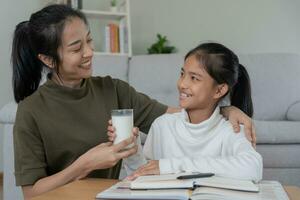 asiatisk ung liten flicka lära sig på Hem. do läxa med snäll mor hjälp, uppmuntra för examen. mamma passera på en glas av mjölk till dotter. flicka Lycklig hemskola. mamma lära och råd utbildning tillsammans foto