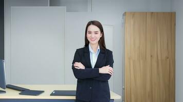 asiatisk professionell arbetssätt kvinna vem har lång hår bär svart formell kostym med blå skjorta medan hon ärm korsning och självsäker ler i kontor rum. foto