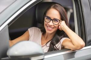 leende brunett med glasögon enheter en bil, med ett ärm ut av de öppen fönster på de sida av henne bil foto