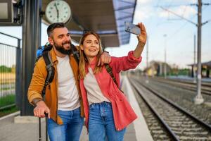 Lycklig par är stående på järnväg station och väntar för ankomst av deras tåg. de är tar selfie. foto
