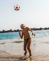 ung pojke spelar med en fotboll boll på de strand förbi de hav foto