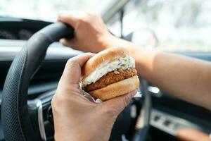 asiatisk lady innehav hamburgare till äta i bil, farlig och risk ett olycka. foto