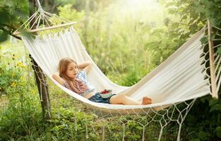 en liten flicka vilar i en hängmatta och äter körsbär i de sommar. foto