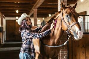 Söt cowgirl kammar henne brun måla häst, leende, stående inuti de stabil foto