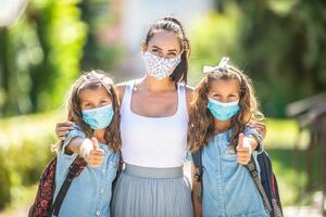 en mor och henne tvilling dotter använda sig av en skyddande mask när återvändande till skola under de covid-19 karantän foto