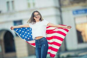 Lycklig ung amerikan skola flicka innehav och vinka i de stad med USA flagga foto
