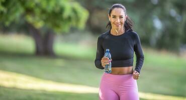 attraktiv ung kvinna löpning i de parkera med flaska av vatten foto