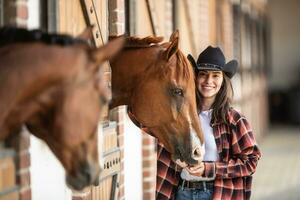 snygg flicka i cowgirl hatt står Nästa till hästar inuti de stabil, leende foto