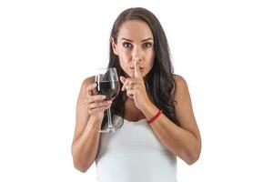 ung kvinna innehav en glas av vin medan innehav henne index finger i främre av henne mun. isolerat bakgrund foto