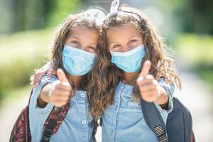 tvilling systrar med ansikte masker gå tillbaka till skola under de covid-19 karantän och som visar tummen upp foto