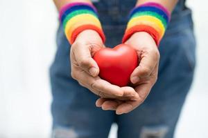 asiatisk dam som bär regnbågsflaggarmband och håller rött hjärta, symbol för HBT stolthet månad firar årliga i juni sociala av homosexuella, lesbiska, bisexuella, transpersoner, mänskliga rättigheter. foto