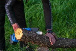 handhållna bärbar camping elektrisk fick syn på på en batteri för skärande ved och trä. närbild, en hand sågning en logga, stickor flygande foto