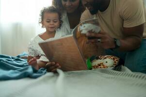 Lycklig afrikansk amerikan föräldrar med liten döttrar spelar , läsning bok på de säng i sovrum på Hem, Lycklig familj begrepp foto