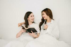 attraktiv lesbisk par använda sig av mobil telefon, läsplatta Kolla på film på säng. skön syskon i pyjamas liggande ner med filt i sovrum tillsammans, i morgon- foto