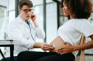 gravid afrikansk kvinna har utnämning med läkare på klinik. manlig gynekolog ob gyn läkare specialist med stetoskop lyssnar till bebis hjärtslag i mors mage. graviditet, hälsa vård foto