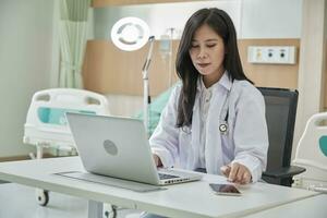 ung skön asiatisk kvinna läkare uppkopplad arbetssätt med bärbar dator och smartphone på medicinsk samråd Ansökan via internet på arbetssätt skrivbord i sjukhus klinik. foto