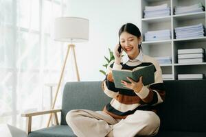 självsäker asiatisk kvinna med en leende stående innehav anteckningsblock och läsplatta i levande rum på de soffa på Hem foto