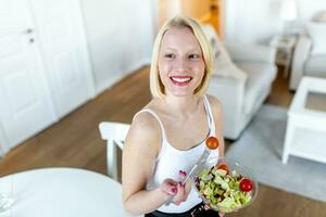porträtt av en Lycklig lekfull flicka äter färsk sallad från en skål i henne kök. skön passa kvinna äter friska sallad efter kondition träna foto