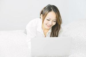 asiatisk kvinna som använder bärbar dator i emotionellt leende medan hon lägger på sängen. livsstil, avkoppling, fritid, teknik kommunikation koncept.