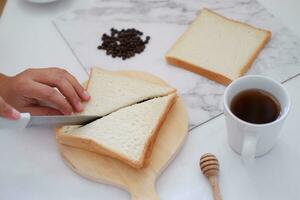 kvinna skärande bröd med kniv och kopp av kaffe på vit tabell. foto