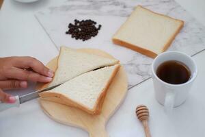 kvinna skärande bröd med kniv och kopp av kaffe på vit tabell. foto