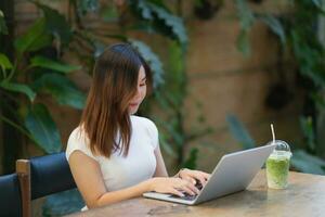 Asiatisk ung kvinna i smarta fritidskläder som arbetar på bärbar dator medan du sitter på kreativa kontor eller café. ung flicka som arbetar med laptop på träbordet i caféet foto