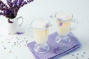 kaffe med mjölk, skum och lavendel- i glasögon och en bukett av blommor i en kanna på en vit tabell. kopia Plats foto
