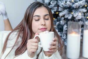 kvinna i värma vit vinter- Tröja liggande i säng på Hem på jul eve innehav kopp med marshmallows, gran träd Bakom foto