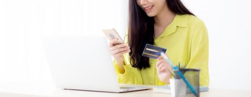 ung asiatisk affärskvinna som använder den smarta telefonen och innehar kreditkortet medan online-shopping och betalning online med bärbar dator på skrivbordet hemma, kvinnligt innehavbetalkort, kommunikationskoncept.
