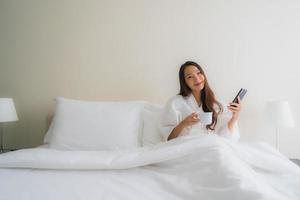 stående vackra unga asiatiska kvinnor med kaffekoppen och mobiltelefonen på sängen foto