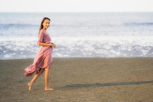 porträtt ung vacker asiatisk kvinna gå leende och glad på stranden havet och havet foto