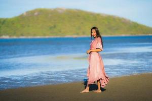 porträtt ung vacker asiatisk kvinna gå leende och glad på stranden havet och havet foto