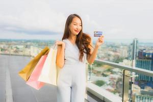 stående vacker ung asiatisk kvinna glad och ler med kreditkort för shoppingväska från varuhuset foto