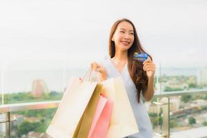 stående vacker ung asiatisk kvinna glad och ler med kreditkort för shoppingväska från varuhuset foto