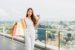 stående vacker ung asiatisk kvinna glad och ler med kreditkort för shoppingväska från varuhuset foto