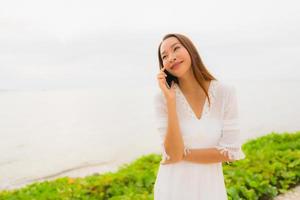 porträtt vacker asiatisk kvinna slitage hatt med leende glad för att prata mobiltelefon på stranden foto