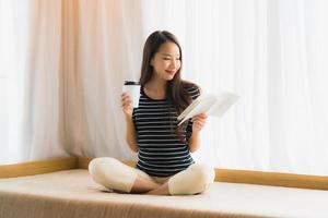 porträtt vacker ung asiatisk kvinna läsebok och hålla kaffekopp eller mugg i soffan i vardagsrummet foto