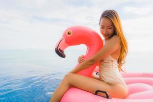stående ung asiatisk kvinna på uppblåsbar flottörflamingo runt utomhuspool i hotellresort foto